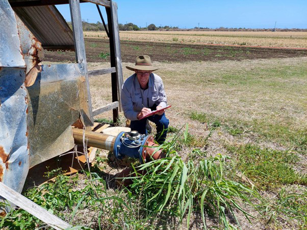 A white male farmer kneeling next to a rural water meter, with a metal structure to his right and broadacre farmland in the background
